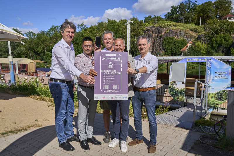 Matthias Lux und Peter Kolbert (beide Stadtwerke GmbH) halten gemeinsam, mit  Mark Lange vom Stadtmarketing Halle, Andreas Blümner vom Smart-City-Team Halle (Saale) und David Hauff von der Wassertouristik Saaletal, eine Werbetafel in der Hand.