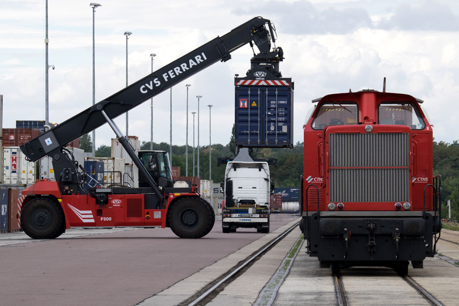 Am rechtsen Bildrand steht eine tote Lok, die frontal fotografiert wird. Von links kommt eine Art übergroßer Stapler, an dem ein dunkelblauer Container hängt.