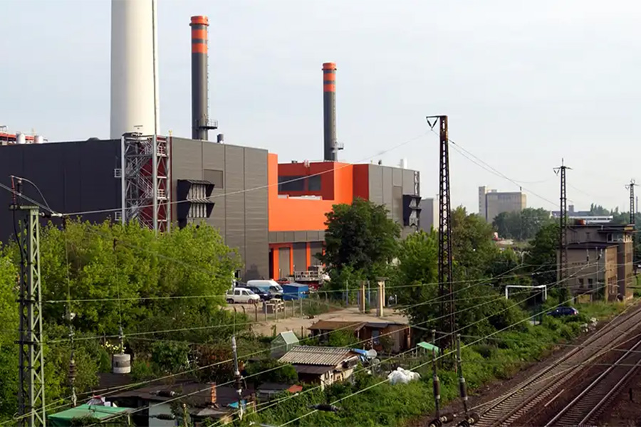 Kraftwerk mit orange-grauer Fassade und zwei Schornsteinen, davor Bahngleise und Oberleitungen.