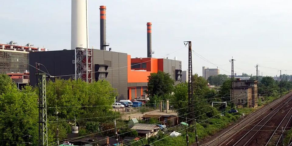 Kraftwerk mit orange-grauer Fassade und zwei Schornsteinen, davor Bahngleise und Oberleitungen.